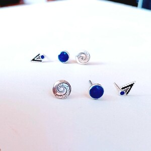 Op de afbeelding: Een verzameling van zes oorknopjes met drie verschillende ontwerpen: twee zilveren driehoekige knopjes met blauwe accenten, twee ronde knopjes met een diepblauw midden en twee zilveren ronde knopjes met een gestructureerd spiraalpatroon.