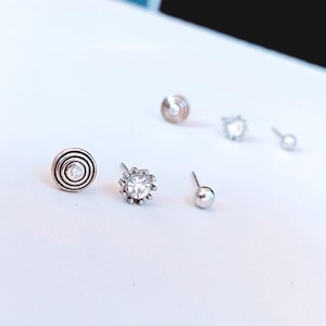 Peut inclure: Un ensemble de cinq boucles d'oreilles à tige argentées. Le lot comprend une boucle circulaire avec des anneaux concentriques et une pierre centrale, une boucle en forme de fleur avec une pierre claire, deux billes lisses et un petit clou en cristal.