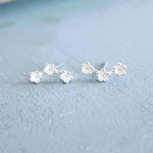 Peut inclure: Une paire de boucles d'oreilles à tige argentées, chacune composée de trois petites fleurs texturées disposées en arc de cercle. Le métal présente une finition brillante et réfléchissante, et chaque boucle d'oreille est dotée d'une tige visible.