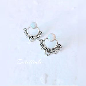May include: A pair of silver-toned earrings with a white, opal-like stone at the top and a decorative, floral-inspired design. The earrings have a circular base and are set against a white background. The text "Detellinka" is visible.