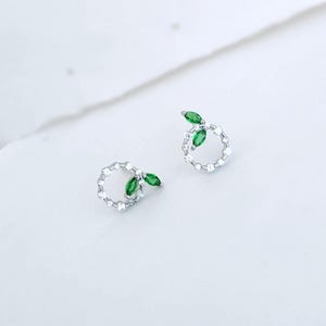 Peut inclure: Une paire de boucles d'oreilles en argent. Chaque boucle d'oreille présente un motif circulaire incrusté de petits cristaux transparents. Un groupe de trois pierres vertes taillées en marquise est fixé à chaque cercle, ressemblant à des feuilles.