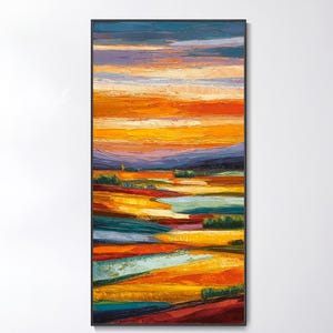 Puede incluir: Pintura abstracta de paisaje con marco negro. La obra presenta bandas horizontales de color, incluyendo naranja, amarillo, azul y rojo, evocando una puesta de sol sobre un campo. La pintura se exhibe sobre un sofá blanco.