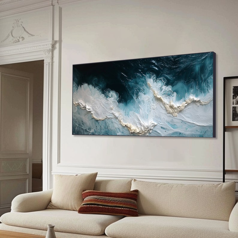 May include: Abstract painting of ocean waves in shades of blue and white. The painting is framed and hanging on a white wall above a beige couch.