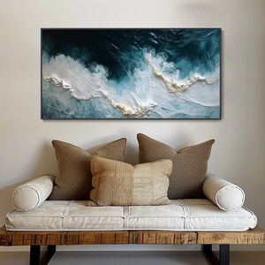 May include: A white and blue abstract painting of ocean waves with gold accents. The painting is framed in black and hanging on a white wall above a white couch with brown pillows.