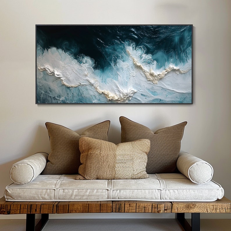 May include: A white and blue abstract painting of ocean waves with gold accents. The painting is framed in black and hanging on a white wall above a white couch with brown pillows.
