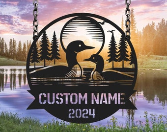 May include: Black metal sign with a silhouette of two loons, trees, and a sun. The sign has a banner with the text "CUSTOM NAME 2024".