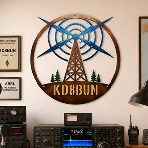 Puede incluir: Un letrero de pared circular de metal con la silueta de una torre de radio marrón, detalles de rayos azules y pinos verdes. El texto KD8BUN se muestra en letras oscuras y negritas en la parte inferior del marco circular, centrado en la pared.