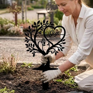 Könnte beinhalten: Ein schwarzer Metall-Gartenstecker in Form eines Baumes mit einem herzförmigen Ausschnitt. Das Herz enthält den Namen "Gigi" und die Daten "2012-2024". Ein Schmetterling ist ebenfalls im Design enthalten. Der Stecker wird in die Erde gesteckt.