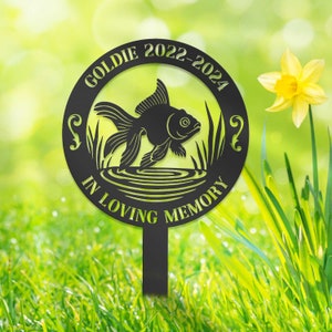 Puede incluir: Estaca de jardín de metal negro con diseño de pez dorado. La estaca tiene el nombre "Goldie" y los años "2022-2024" inscritos, junto con las palabras "In Loving Memory". El pez dorado está rodeado de plantas acuáticas.