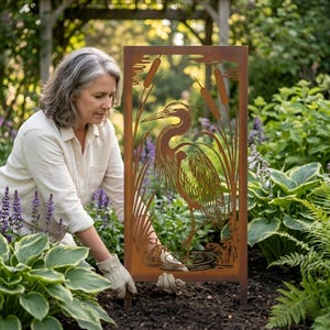 May include: A rectangular, rust-colored metal garden art piece featuring a heron standing in water, surrounded by cattails and reeds. The heron is intricately cut out of the metal. The art is placed in a garden setting with lush greenery and purple flowers.