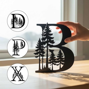 May include: A black metal letter "B" with a forest scene cut out. The letter is being held up by a hand. Three circular designs with the letters "D", "P", and "X" and forest scenes are also visible.