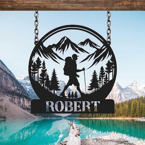May include: Black metal silhouette of a hiker with a backpack walking through a mountain landscape. The silhouette is in a circle with the name "ROBERT" below it.