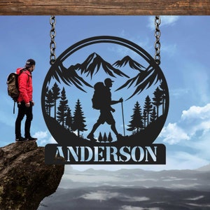 May include: Black metal silhouette of a hiker with a backpack walking through a mountain landscape with trees. The silhouette is in a circle with the name "ANDERSON" below it.