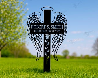 May include: Black metal memorial cross with angel wings, a halo, and a rose. The cross features the name "ROBERT S. SMITH" and dates "09.20.1962-06.15.2025". The cross is set in a grassy field with a blurred background of trees and sky.
