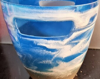Puede incluir: Una maceta de resina azul y blanca con un diseño de playa. La maceta tiene un asa recortada y un fondo arenoso.