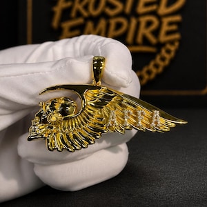 May include: A gold-toned pendant featuring a skull integrated with a large, detailed wing. The wing surface displays the letters AFFA accented with small, clear stones. The pendant includes a top bail for attachment to a chain or necklace.