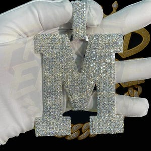 Puede incluir: Un colgante con forma de la letra "M" incrustado con diamantes. El colgante plateado está cubierto de pequeñas piedras brillantes. Se ve una cadena dorada en la parte inferior. El colgante es sostenido por una mano enguantada sobre un fondo negro.
