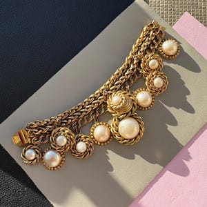 May include: A vintage gold-tone necklace with a chunky chain and multiple pearl and gold-tone pendant charms. The necklace is displayed on a gray surface with a black and pink background.