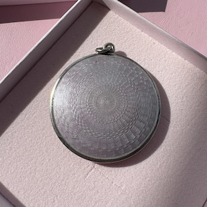 Op de afbeelding: Een cirkelvormige, zilverkleurige hanger met een gedetailleerd, concentrisch guilloché-patroon dat vanuit het midden uitstraalt. Het metalen oppervlak heeft een metaalachtige glans en een klein, geïntegreerd oogje aan de bovenkant voor bevestiging.