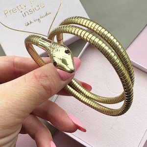 Könnte beinhalten: Ein goldfarbenes Schlangenarmband mit einem detaillierten Schlangenkopf mit roten Edelsteinaugen. Das Armband wickelt sich um das Handgelenk und zeigt ein strukturiertes, segmentiertes Design. Der Hintergrund zeigt eine rosa Schachtel mit dem Text "Pretty Inside" und "shh by sadie."