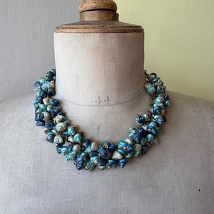 May include: A statement necklace featuring a dense cluster of small, spiraled seashells in varying shades of deep navy blue, turquoise, and iridescent white. The shells are arranged in a thick, rounded collar shape, creating a textured appearance.