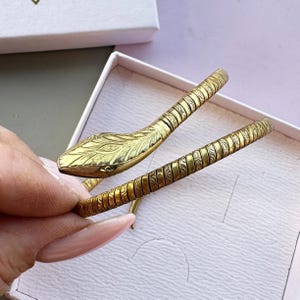 May include: A gold-toned snake-shaped bracelet featuring a textured, segmented body with intricate engraved patterns. The head of the snake is detailed with etched scales and eye markings, while the flexible, coiled design wraps around the wrist.