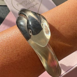 May include: A translucent, smoke-colored resin bracelet shaped like a coiled snake. The design features a smooth, polished surface with a gradient effect, transitioning from a darker, opaque charcoal tone at the head to a lighter, clear finish.