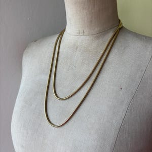 May include: A gold-toned snake chain necklace featuring a smooth, flexible, and rounded design. The necklace is layered, creating two distinct loops of chain that drape evenly across the chest area of the display form in a simple, clean style.