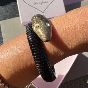 May include: A coiled snake bracelet featuring a textured black body and a metallic gold-toned head. The snake head is detailed with engraved scale patterns and two clear, round, faceted stones set as eyes. The design wraps around the wrist.