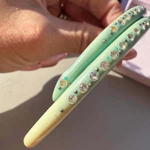 May include: Two stacked bangle bracelets featuring a smooth, glossy finish. The top bangle is a vibrant mint green, while the bottom bangle is a pale cream colour. Both are adorned with a row of embedded, clear, round, faceted crystals.