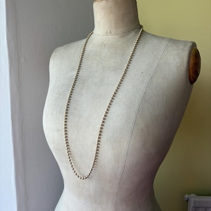 May include: A long, continuous necklace composed of small, uniform, metallic gold-toned spherical beads. The necklace is draped around the neck of a beige fabric-covered mannequin torso, hanging down in a simple, singular loop against the form.