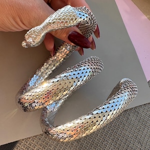 以下が含まれることがあります： とぐろを巻いたヘビの形をしたシルバーカラーの金属製ブレスレット。ヘビの体は柔軟なメッシュ状の質感で、小さな鱗のようなセグメントが重なり合っており、全長にわたって光を反射するきらめく表面を作り出しています。