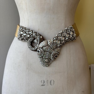 May include: A gold-toned belt featuring a large, central snake-shaped buckle encrusted with clear rhinestones and small white beads. The snake head and coiled body design is densely covered in sparkling crystals, creating a textured, ornate appearance.