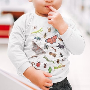 May include: A young child wearing a white long-sleeved shirt with a colourful insect and butterfly print. The child is looking at the camera and has a finger in their mouth.