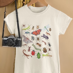 May include: White t-shirt with a colorful insect print. The print features a variety of insects, including butterflies, beetles, ants, and a scorpion.
