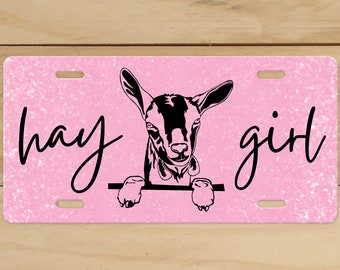 Puede incluir: Placa de matrícula rosa con purpurina con una silueta negra de la cabeza de una cabra asomándose por la parte superior y el texto "hay girl" en cursiva negra.