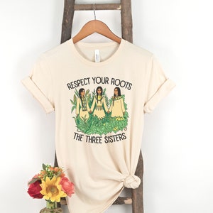 May include: A beige t-shirt with a graphic of three Indigenous women standing in a garden with corn, beans, and squash. The text "Respect Your Roots The Three Sisters" is printed on the shirt.