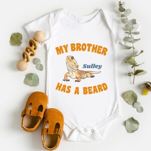 May include: White baby onesie with orange text that reads "My brother Sully has a beard". A cartoon bearded dragon is pictured above the text.