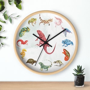 May include: A round wall clock with a light wood frame. The clock face features watercolor illustrations of various reptiles and amphibians, including a lizard, chameleon, and frog. Black clock hands indicate the time.