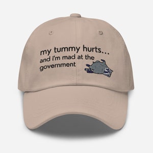 May include: A tan baseball cap with the text "my tummy hurts... and I'm mad at the government" embroidered on the front. A small embroidered raccoon is lying on its back next to the text.