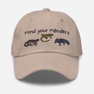 Może przedstawiać: Beżowa czapka z daszkiem z haftowanym czarnym napisem "mind your manders". Na czapce znajdują się trzy małe haftowane obrazki salamander w różnych kolorach.