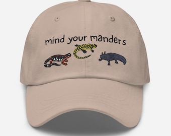 May include: A beige baseball cap with the text "mind your manders" embroidered in black. The cap features three small embroidered images of salamanders in different colors.