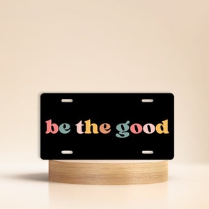 May include: Black license plate with white lettering that reads "be the good" in a colorful retro font.
