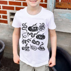 May include: White t-shirt with a black and white print of various snakes. The snakes are arranged in a pattern on the shirt.