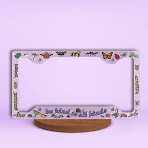 May include: A purple license plate frame with a white background and a variety of colorful insect illustrations. The text "be kind to all kinds" is printed in purple on the frame.