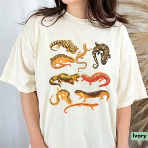 May include: A white t-shirt with a print of various salamanders in different colors and patterns. The salamanders are arranged in a random pattern on the shirt.