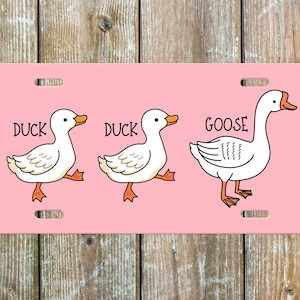 May include: A pink license plate with a white duck, a white duck, and a white goose. The ducks are labeled "DUCK" and the goose is labeled "GOOSE".