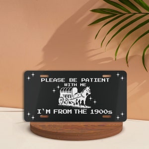 May include: Black metal license plate with white text and a pixelated image of a horse-drawn wagon. The text reads "Please be patient with me I'm from the 1900s".