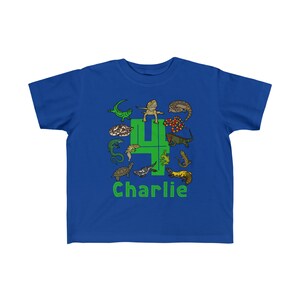 K&ouml;nnte beinhalten: Ein k&ouml;nigsblaues T-Shirt mit einer gr&uuml;nen Zahl vier, die von Comic-Reptilien umgeben ist. Der Text "Charlie" ist unter der Zahl gedruckt.