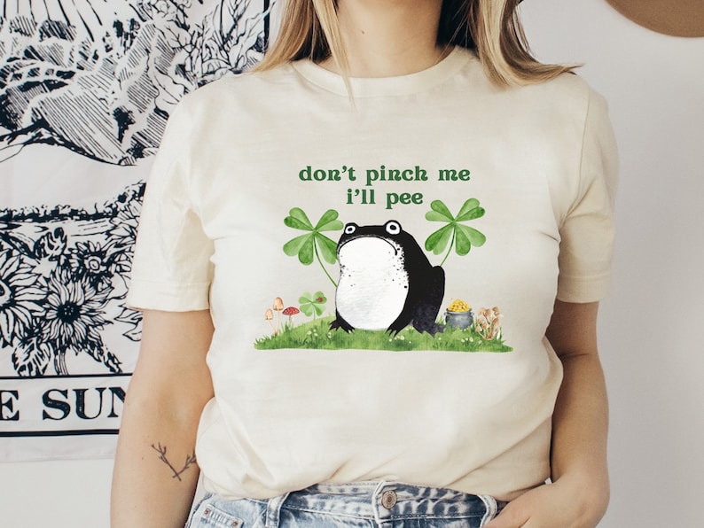 May include: A white t-shirt with a green shamrock and a black frog illustration. The text "don't pinch me I'll pee" is printed on the shirt.